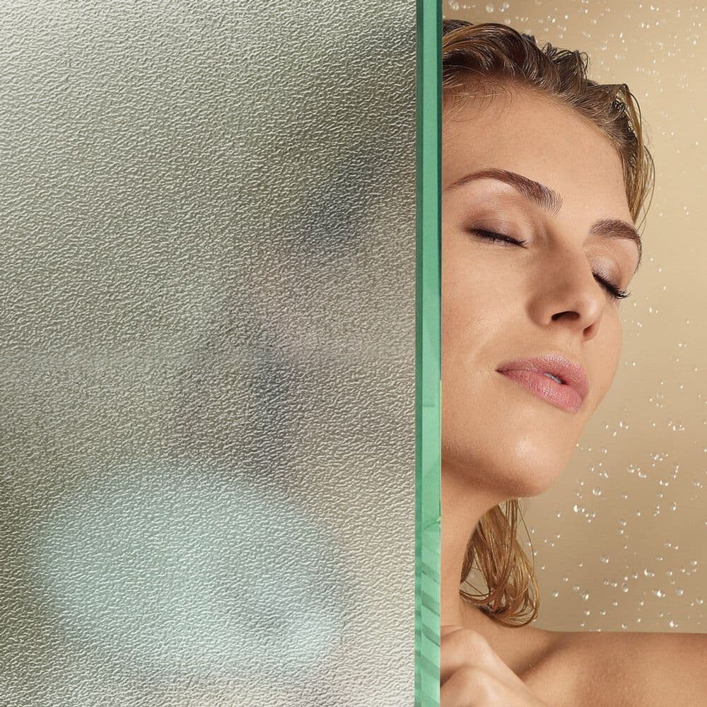 A woman enjoying a shower behind a textured glass shower door, showcasing custom glass designs, including tempered glass and beveled glass edges.