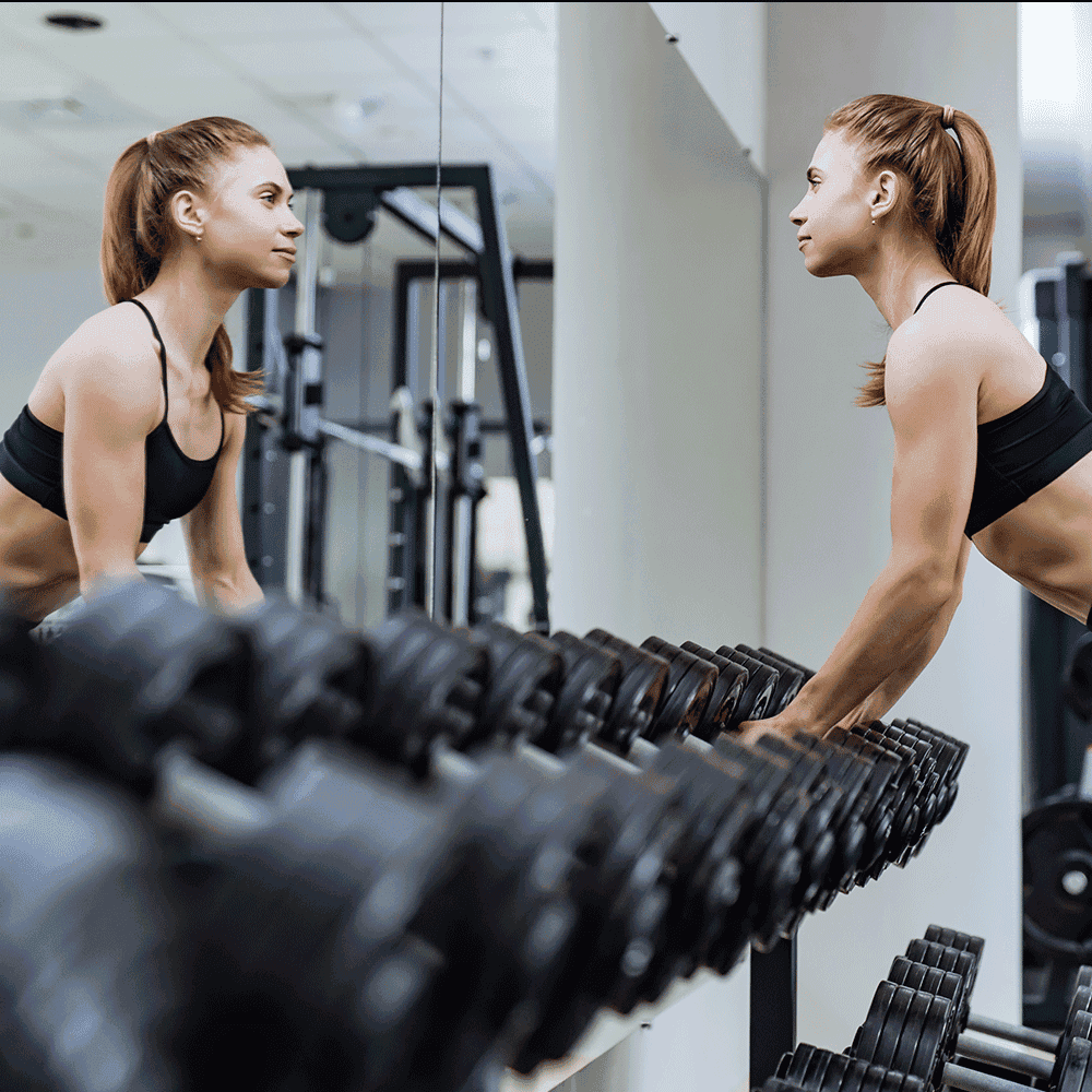 Fit woman exercising in a gym, using free weights, with a large mirror reflecting her form, showcasing a modern fitness environment suitable for custom glass installations like mirrors and beveled glass edges.