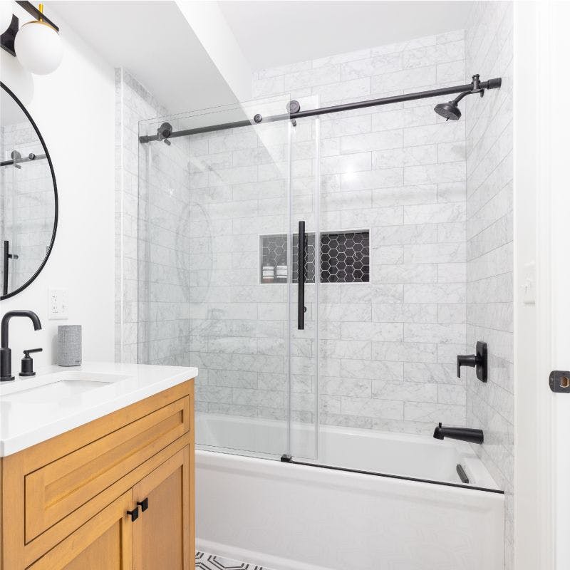 A bathroom with a white marble floor and a stylish black and white tile pattern. Glass sliding doors to bathtub