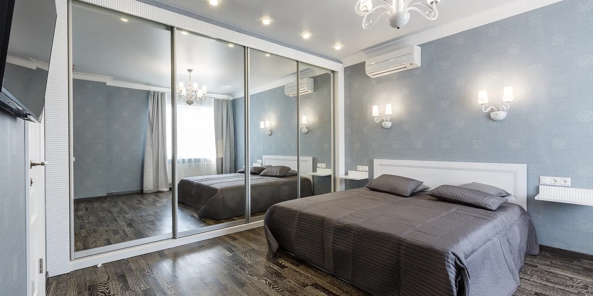 A modern bedroom with light blue wallpaper and a bed with grey linens, featuring a large, floor-to-ceiling sliding mirrored closet.
