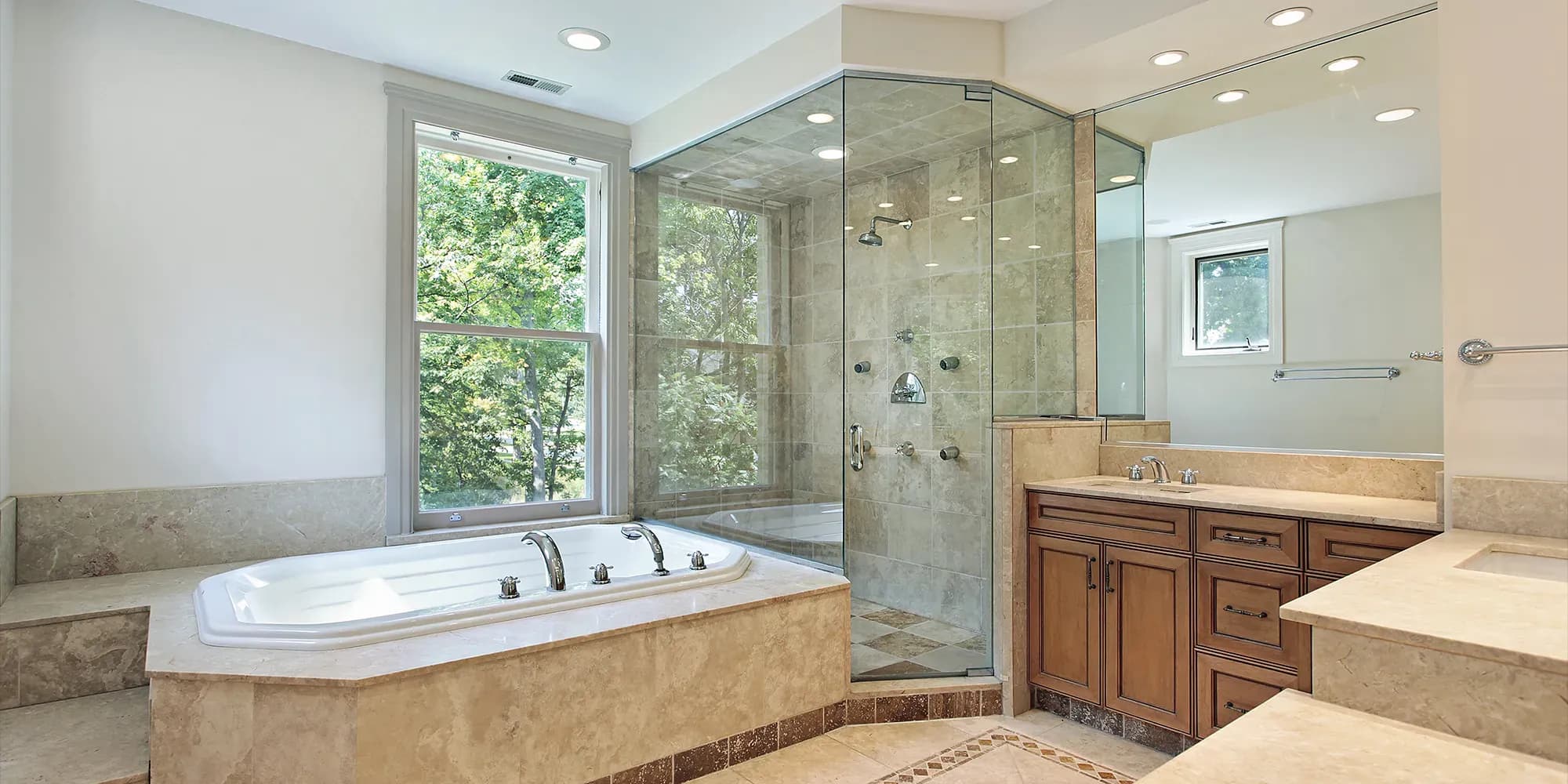 A traditional luxury bathroom with marble tiles, featuring a corner frameless glass shower, a large soaking tub under a window, and a wood vanity.