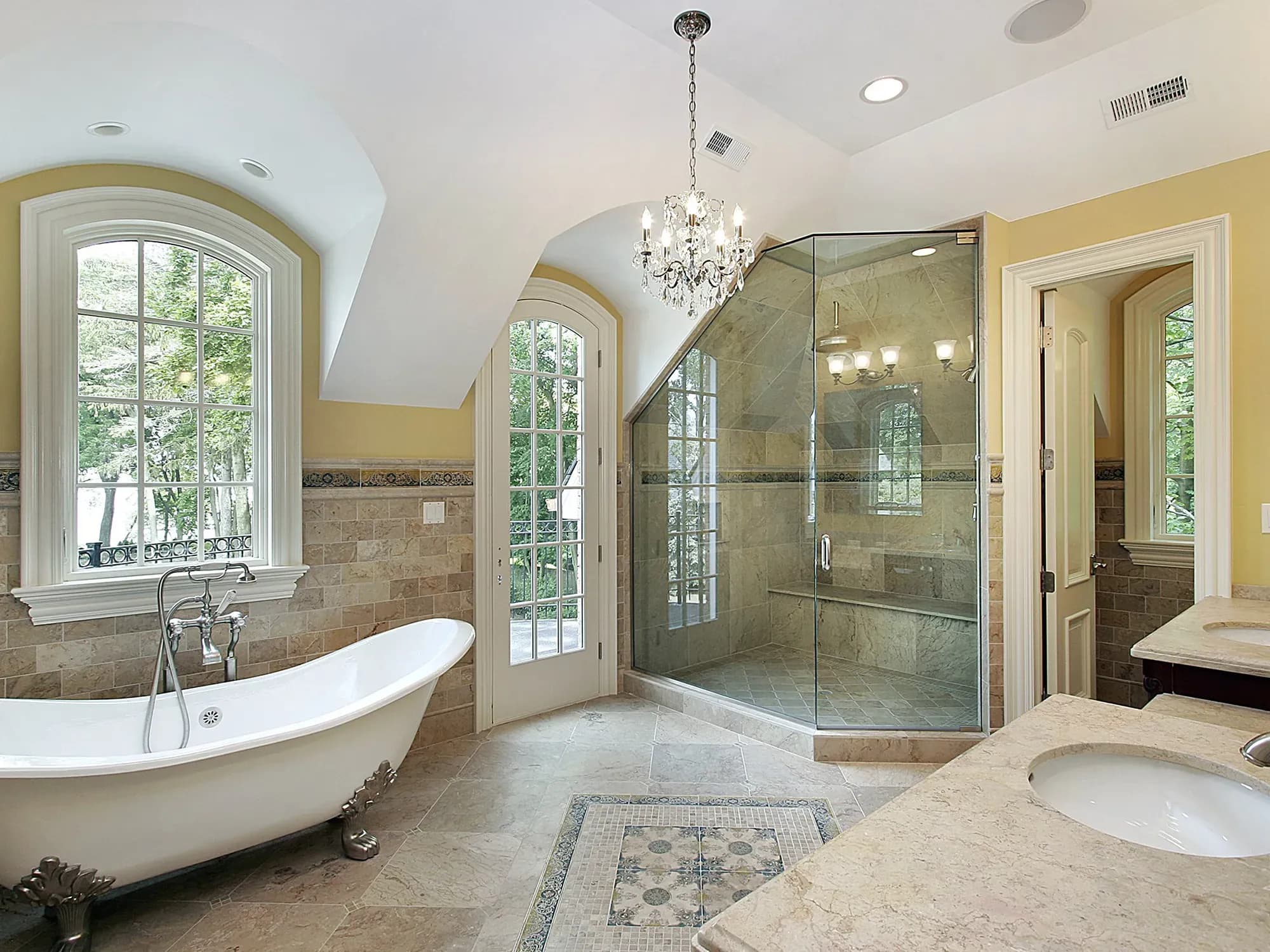 An elegant bathroom with a custom frameless glass shower enclosure fitted under an arched ceiling, a classic clawfoot tub, and travertine tile floors.