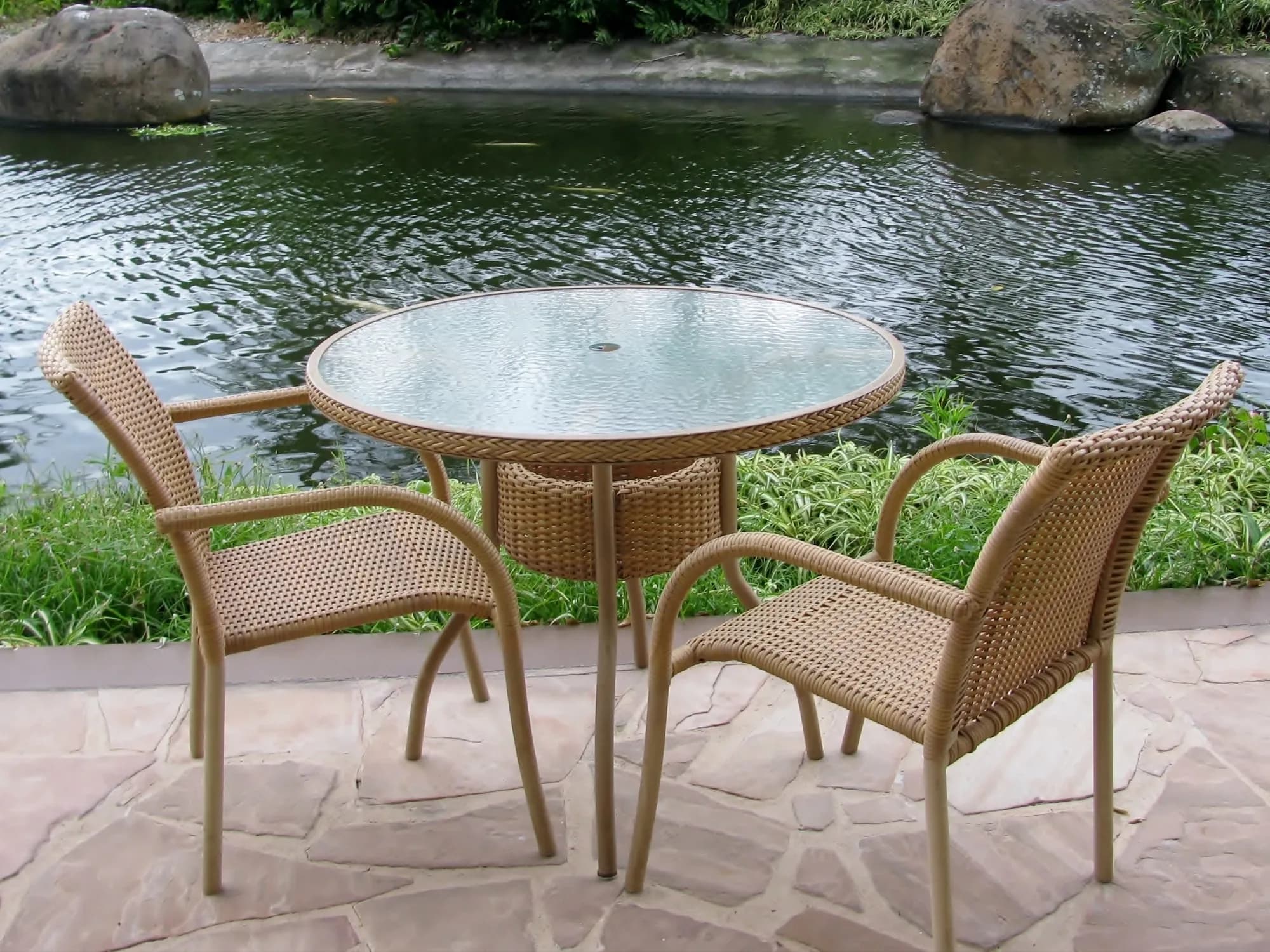 An outdoor wicker patio set with a round, textured glass-top table and two chairs, placed on a stone patio overlooking a tranquil pond.