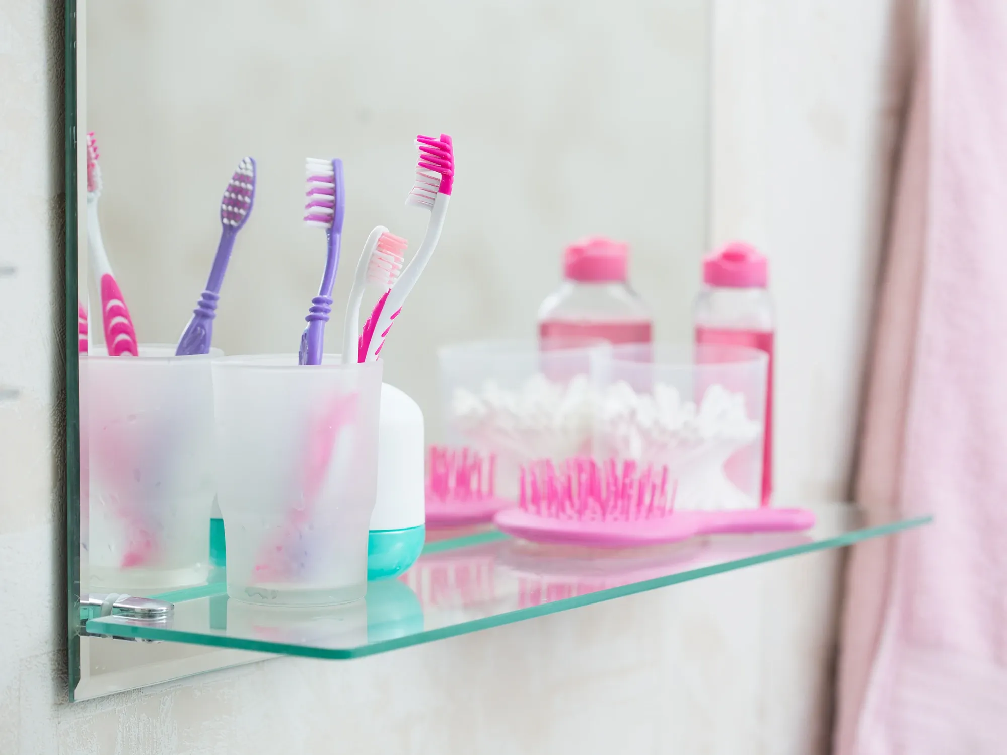 A clear glass bathroom shelf mounted in front of a mirror, holding toothbrushes in frosted cups and other toiletries.