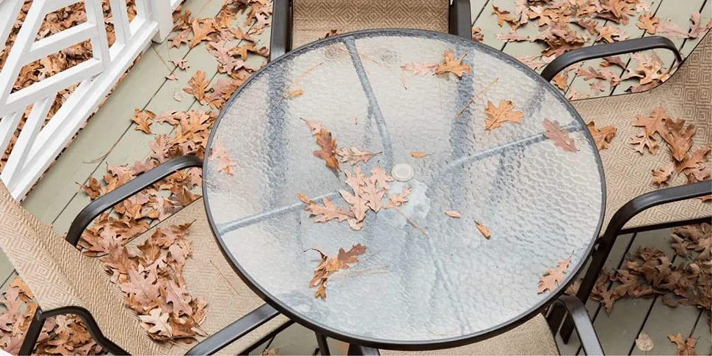 Round glass table top with textured surface covered in fallen leaves, surrounded by chairs on a wooden deck, showcasing custom glass options for outdoor furniture.