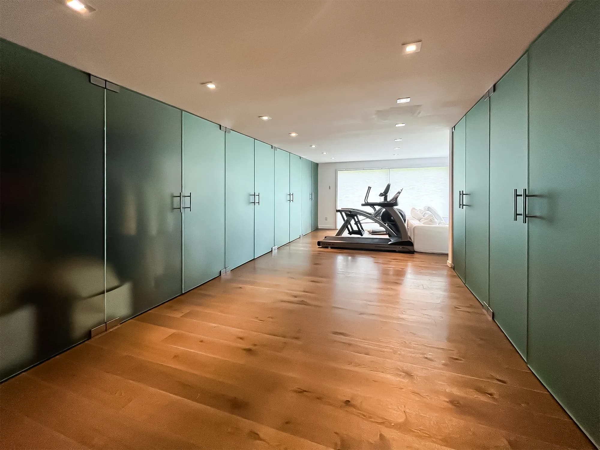 A spacious, modern room with hardwood floors, lined on both sides with floor-to-ceiling frosted glass closets, and a home gym area with a treadmill at the far end.